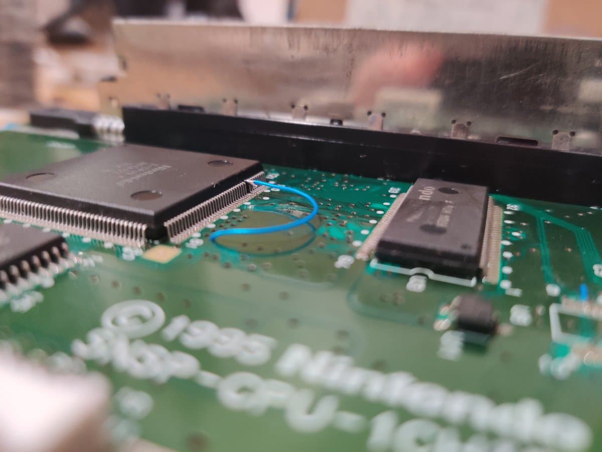 SNES 1-chip motherboard with lifted CPU pins, blue jumper wire, and black heatsink during 50/60Hz mod installation.