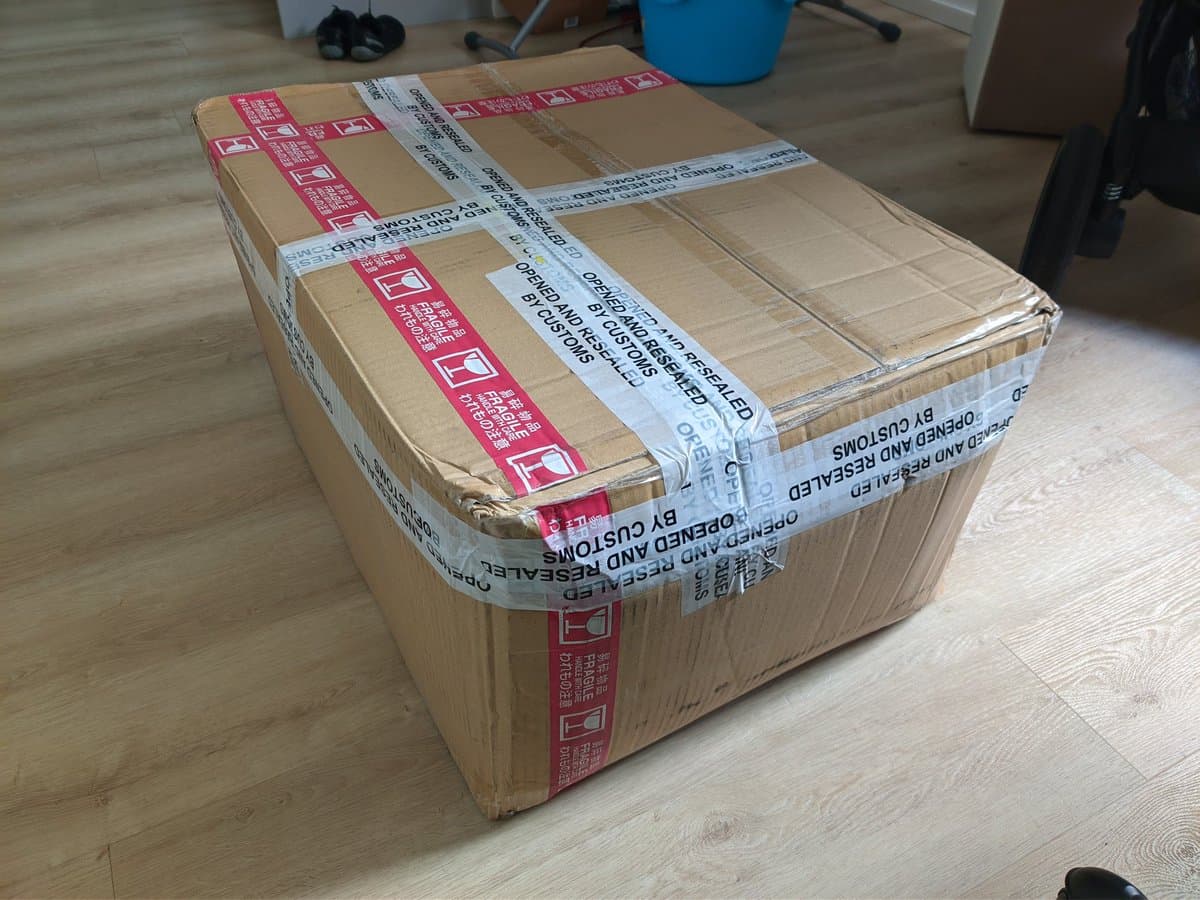 Large cardboard box sealed with red and white warning tape on wooden floor, showing shipping damage from Japan import customs hold.