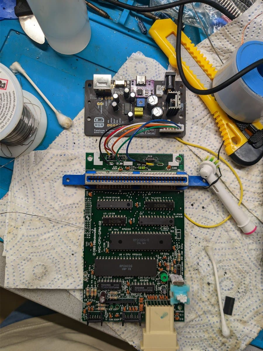 Famicom motherboard with RGB power board installed, composite adapter board with blue bracket, and multicolored wiring connections during repair and modding work.