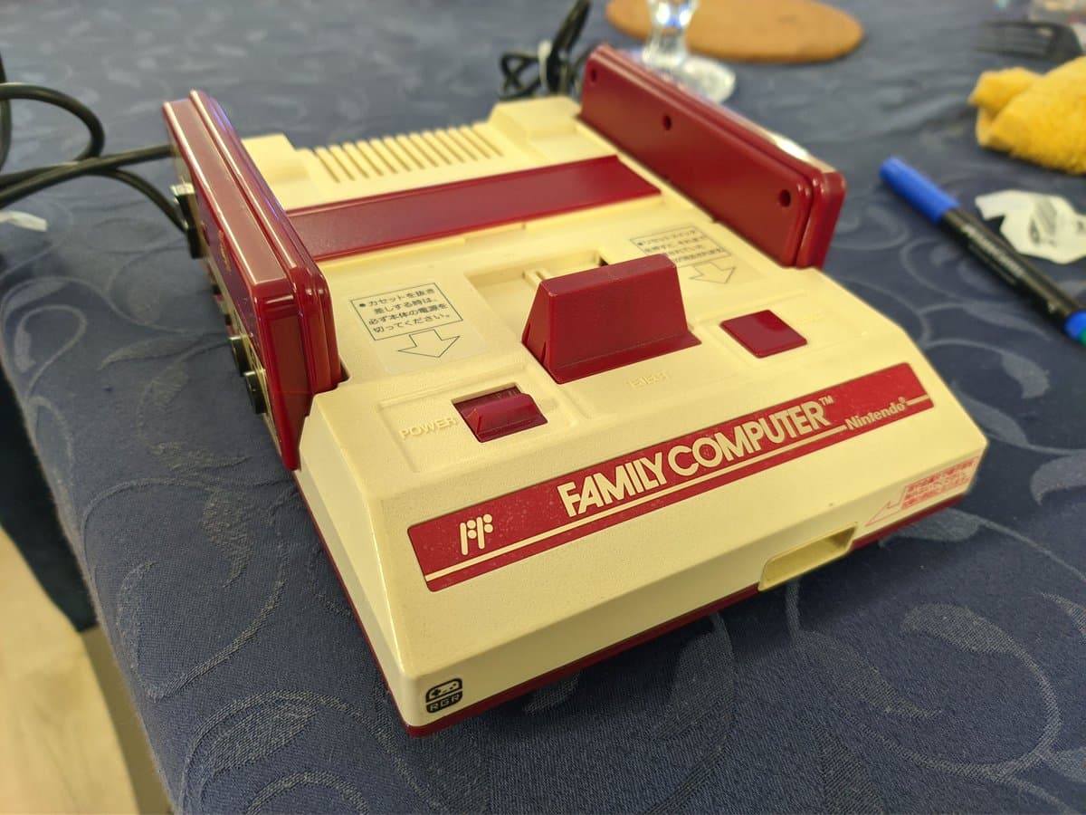 Red and cream Famicom console with controller ports visible, composite mod board installed, new stickers applied, displayed on work surface with tools nearby.