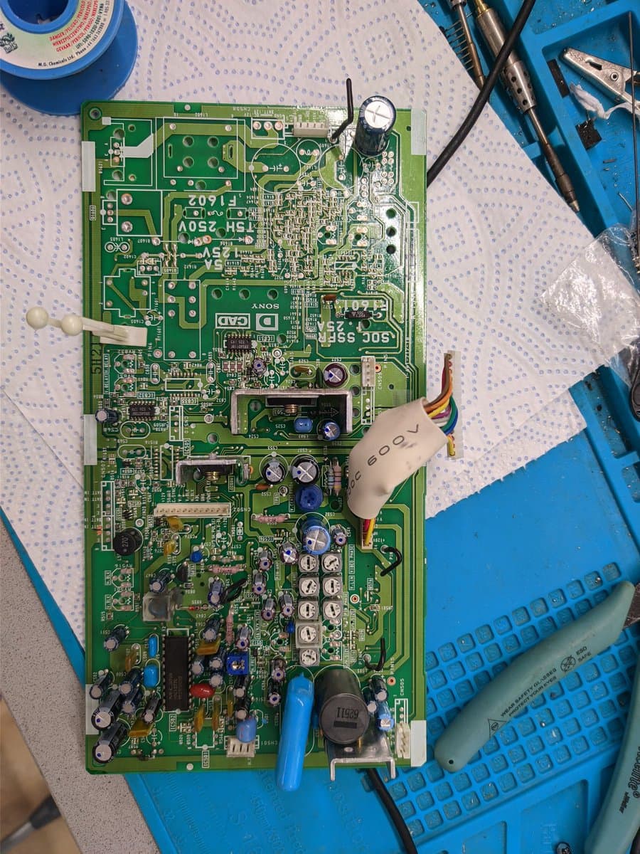Sony PVM-9040 deflection board with capacitors, transistors, and connectors visible; multimeter probe and soldering iron in use during recapping maintenance.