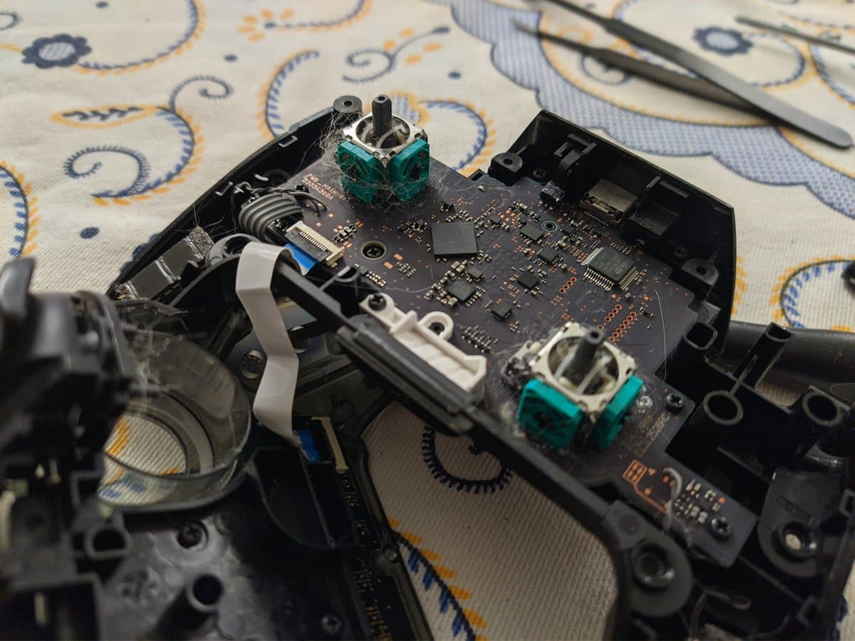 Disassembled Switch Pro controller PCB with teal potentiometers exposed, tweezers visible, showing analog stick module repair work.