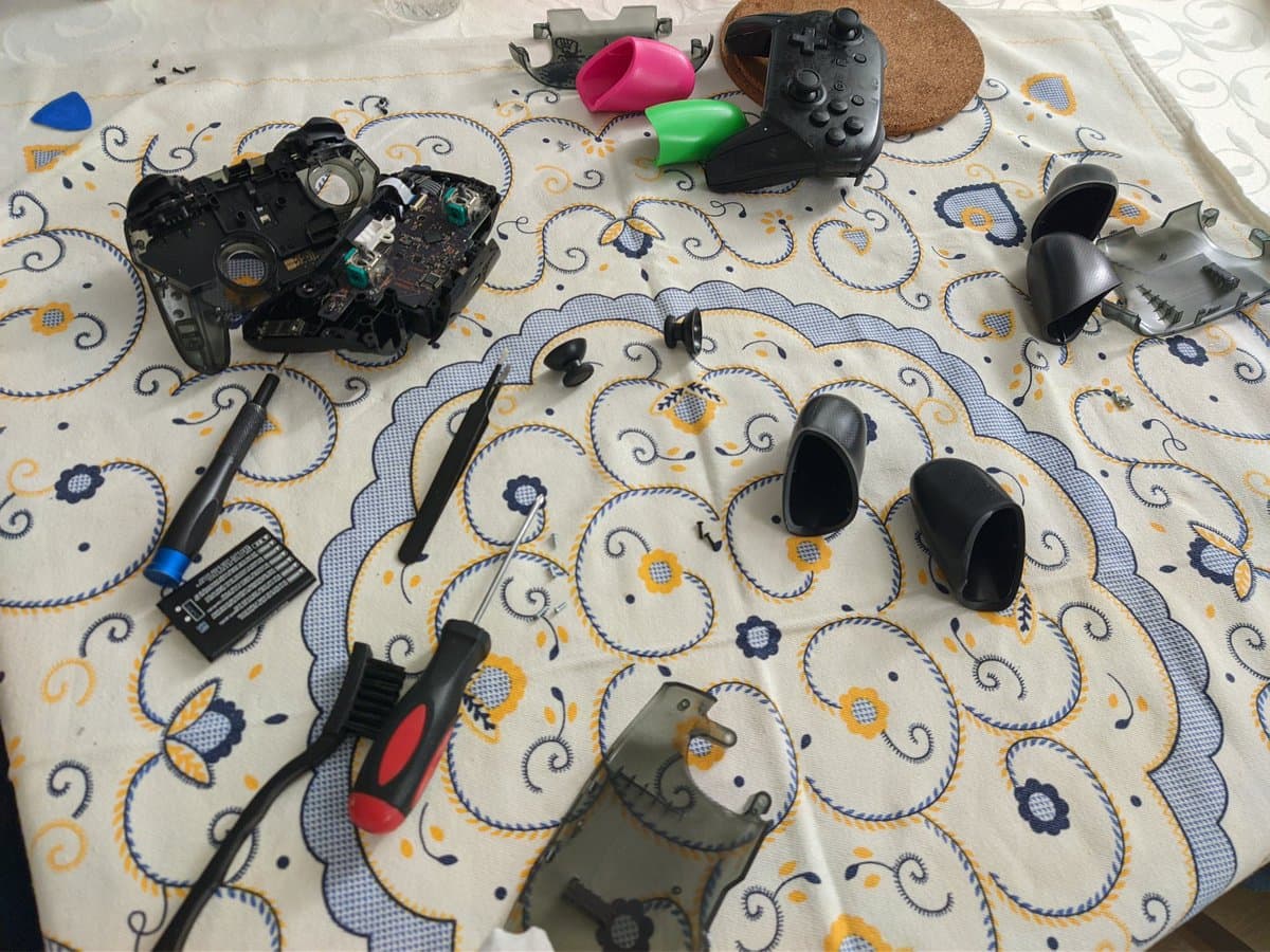 Six disassembled Nintendo Switch Pro controllers with exposed internals, potentiometers, and analog sticks on patterned fabric with cleaning tools and cork mat.