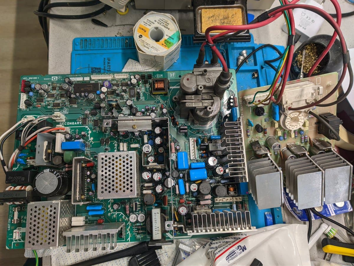 BVM-D14H1E mainboard with capacitors partially replaced; blue solder wick spool and tools visible on blue mat workspace.