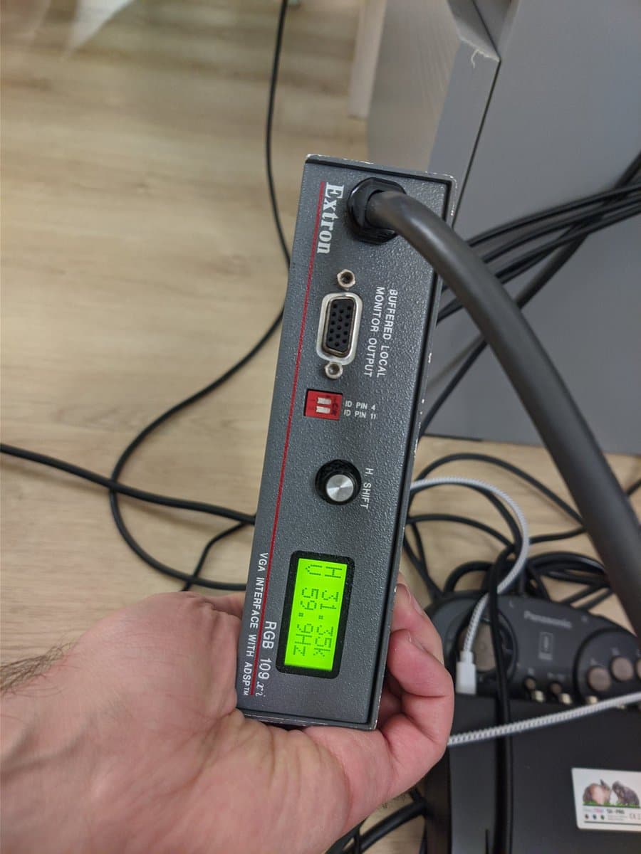 Extron RGB 109xi scaler held in hand, displaying green LCD screen with sync settings, VGA connector visible, cables in background.