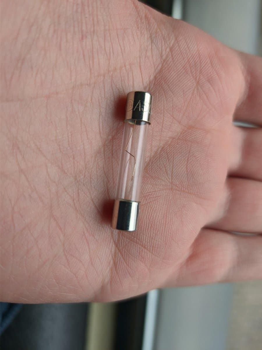 Glass fuse with metal caps held in palm, showing transparent tube interior after mechanical shock damage from monitor collision.