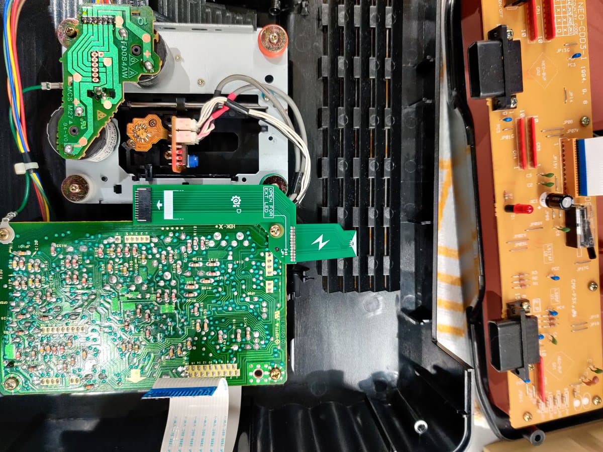 SNK Neo Geo CD motherboard with SD loader mod installed, showing green PCB with components and orange F-RAM upgrade board visible on right side.