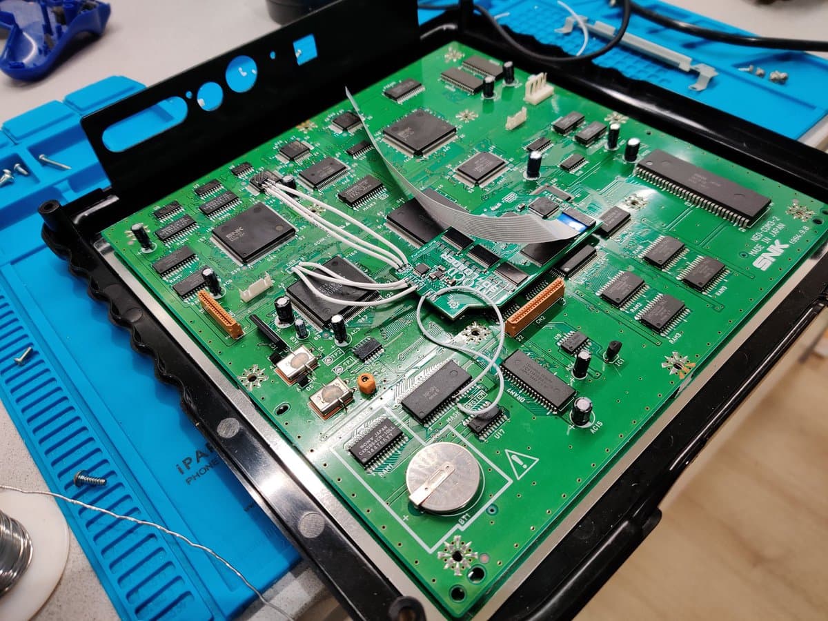 SNK Neo Geo CD mainboard with SD loader and F-RAM upgrade installed, showing integrated circuits, ribbon cables, and coin cell battery replacement mod.