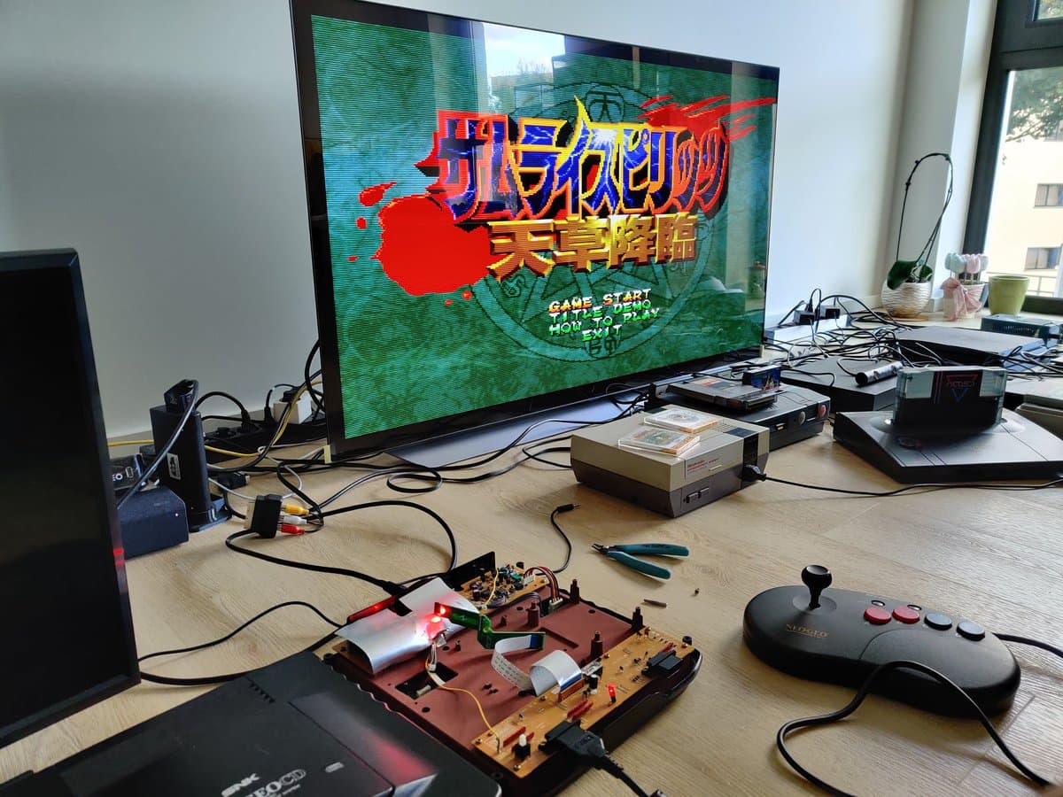 SNK Neo Geo CD with SD loader and F-RAM upgrade installed, displaying title screen on monitor with modded circuit board and controller visible on workbench.