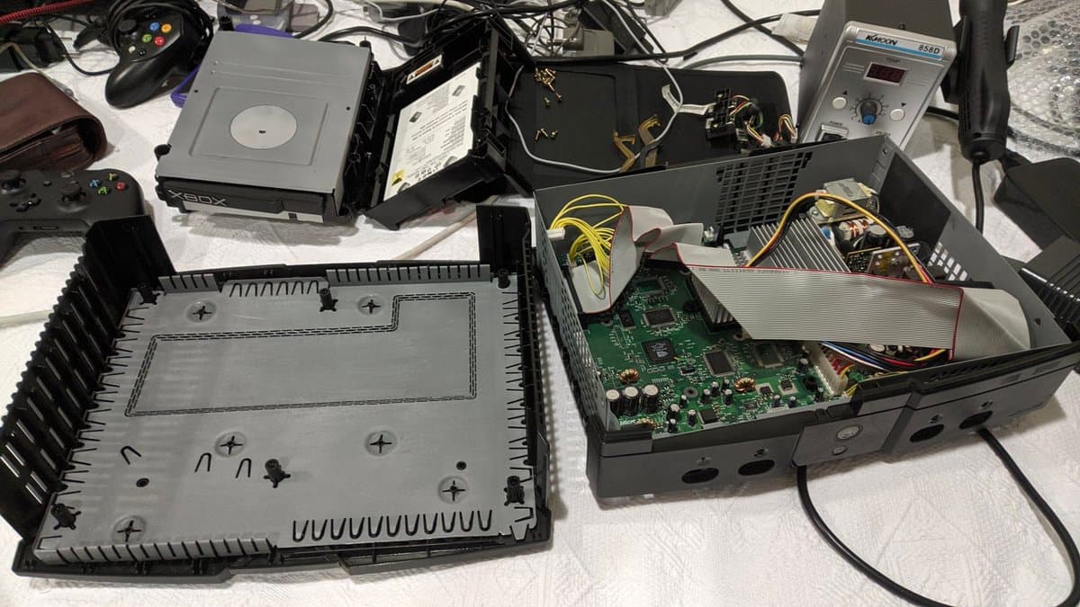 Disassembled OG Xbox showing opened chassis, motherboard with capacitors exposed, and removed bottom panel with heatsink fins visible.