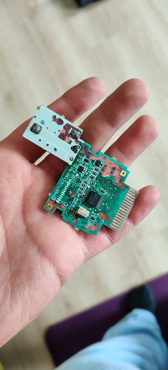 Hand holding repaired N64 Rumble Pak PCB with green circuit board, showing trace repair work and reattached white connector ribbon.