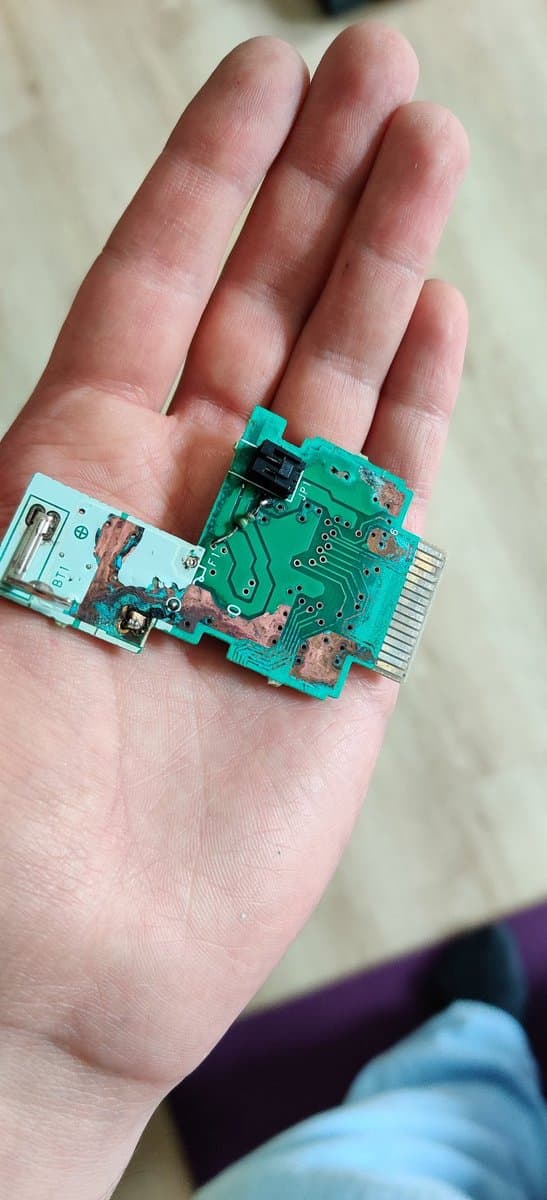 N64 Rumble Pak PCB held in palm showing green circuit board with corrosion damage and repaired traces from battery leak.
