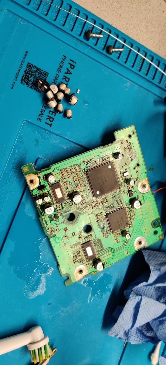 GameCube motherboard with Xeno chip installation in progress, showing three large capacitors removed, PCB components, and brass mounting posts on blue service mat.