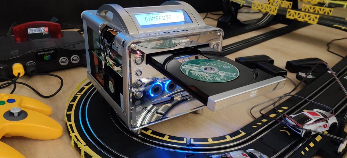 Panasonic Q with open optical drive showing blue LED, disc tray extended with game visible, yellow controller and GameCube console nearby on track racing set.