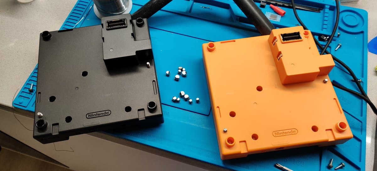 Two recapped Game Boy Players on blue mat: black unit with capacitors nearby, orange unit with reassembled housing during repair work.
