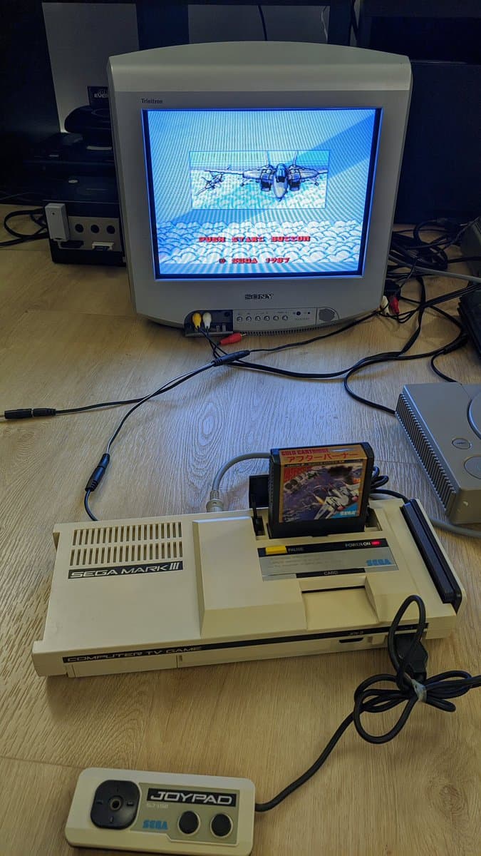 Sega Mark III console connected to CRT monitor displaying Afterburner game, with Joypad controller and game cartridge visible on wooden surface.