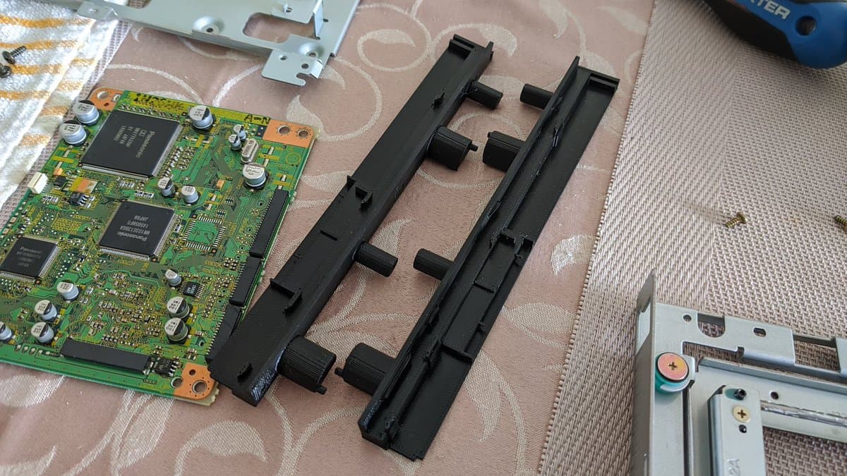 Disassembled Panasonic Q optical drive showing broken black plastic rail alongside 3D printed replacement rails and green circuit board.