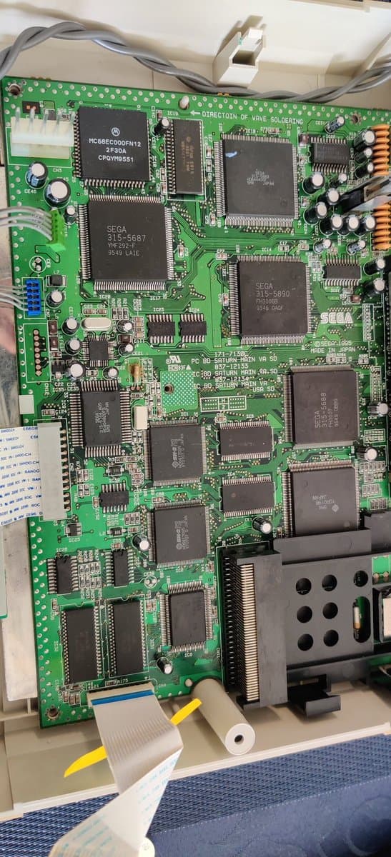 Sega Saturn motherboard with visible IC chips including VDP and sound processors; yellow screwdriver and ribbon cables present during repair work.