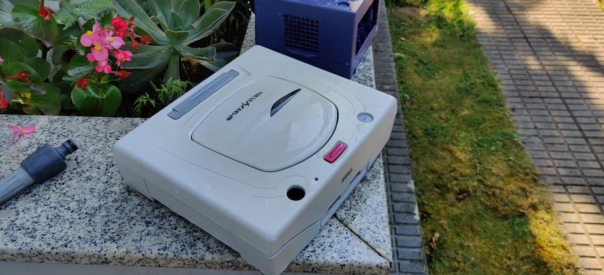 White Nintendo GameCube undergoing retrobrighting treatment outdoors on concrete surface near flowers and moss-covered pathway.