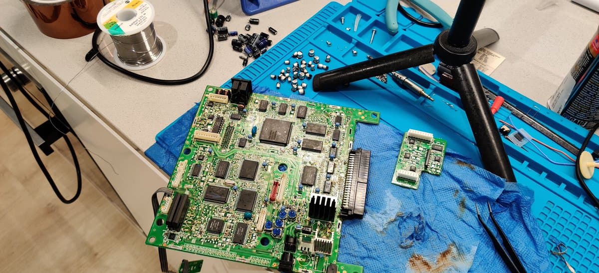 NEC PC Engine Super CD-ROM2 mainboard mid-recap with soldering iron, desoldering braid, and removed capacitors on blue mat.