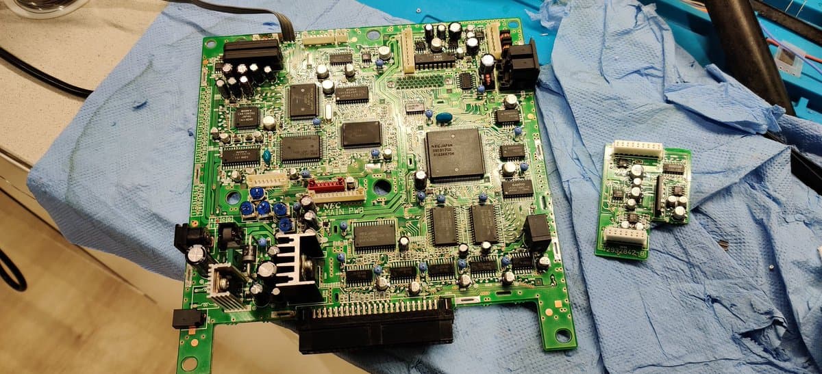 NEC PC Engine Super CD-ROM2 main board mid-recap with removed capacitors on blue paper towel, showing darkened through-holes and flux residue from desoldering work.