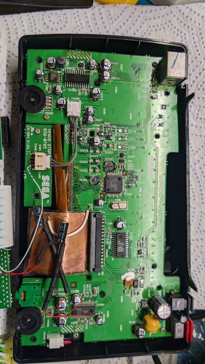 Sega Nomad motherboard with capacitors removed, copper heat sink visible, soldering iron nearby, oxidation and corrosion evident on board surface.