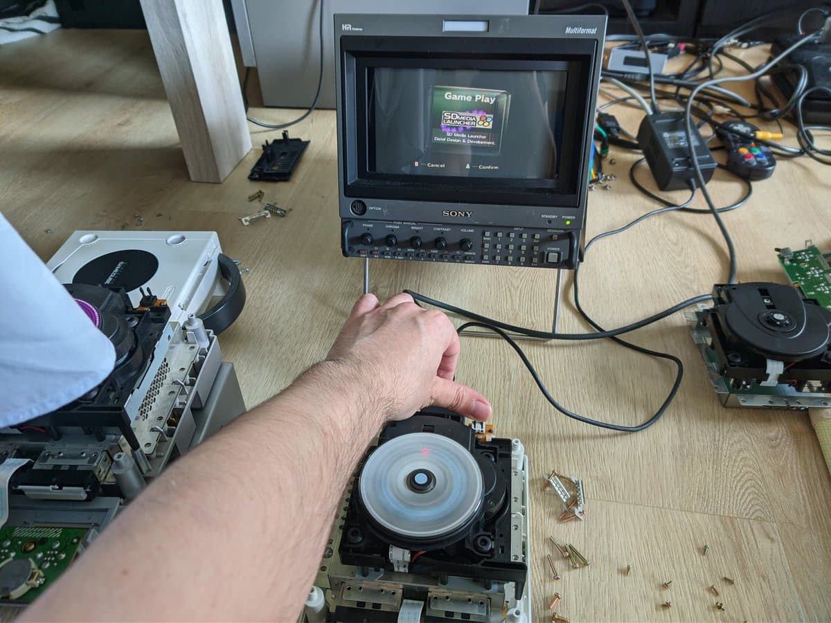 Technician's hand holding GameCube optical drive with recapped capacitors during restoration; Sony monitor displays Game Play menu in background.