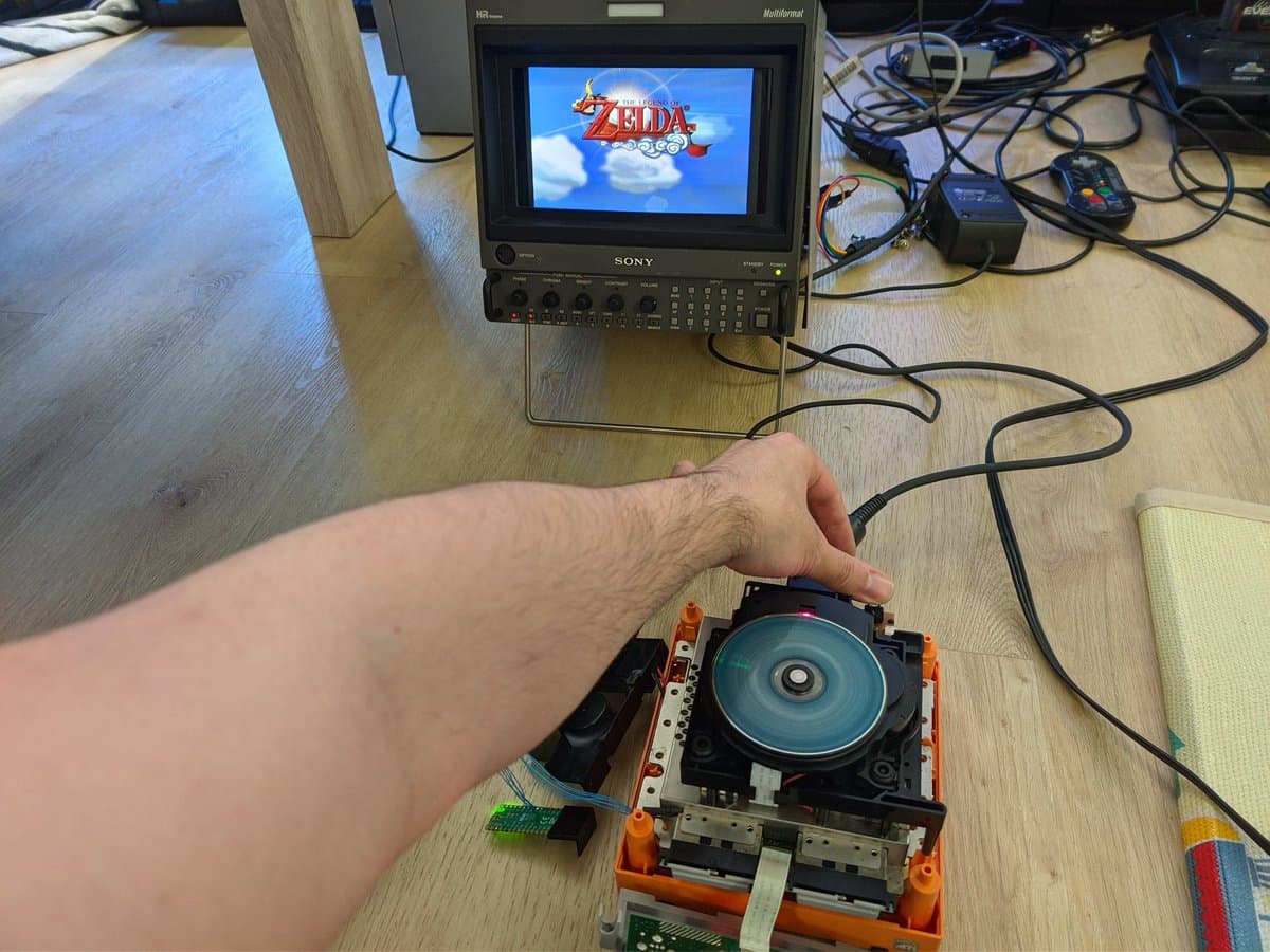 Hand holding open GameCube disc drive showing exposed blue disc, orange housing, and internal components during repair diagnostics on workbench.