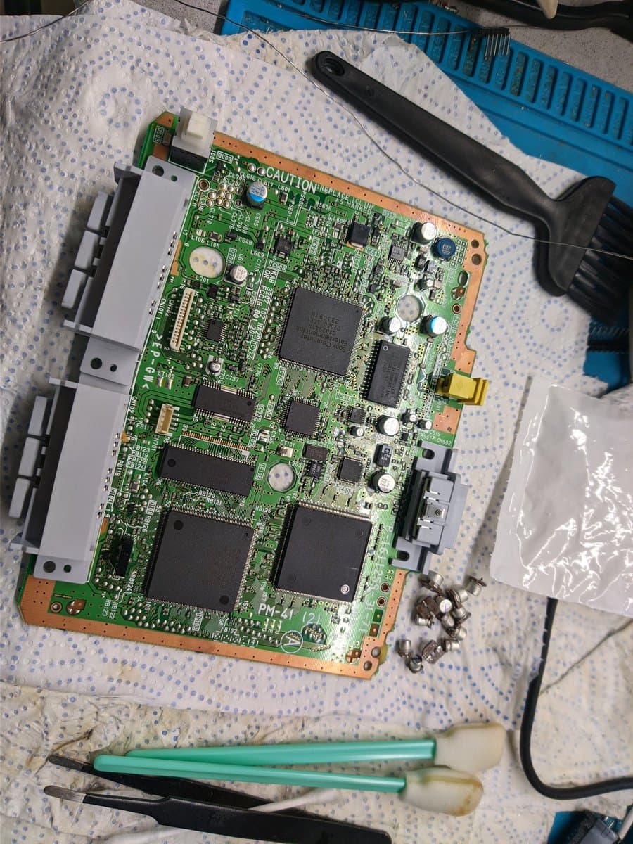 Sony PSOne mainboard during recap repair, SMDs removed, soldering iron and desoldering tools visible on work mat.