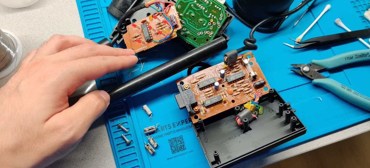 Technician desoldering Power Glove circuit board with iron, orange PCB with exposed components and black connector visible on blue work mat.
