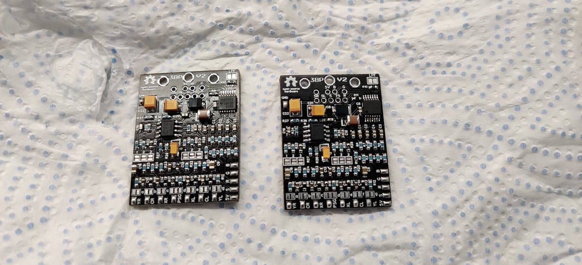 Two assembled 3BP v2 circuit boards side by side on dotted fabric. Gold capacitors and resistors soldered, ICs installed. Clean solder joints visible.