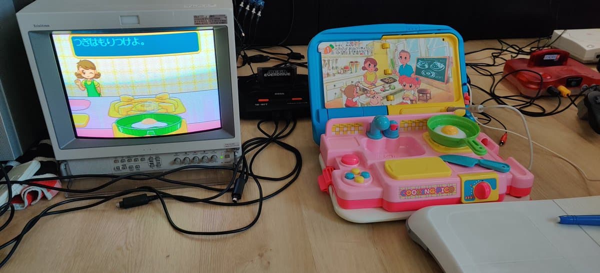 Sega Pico console displaying cooking simulation game on CRT monitor; pink handheld Pico unit with screen showing kitchen scene visible in setup.