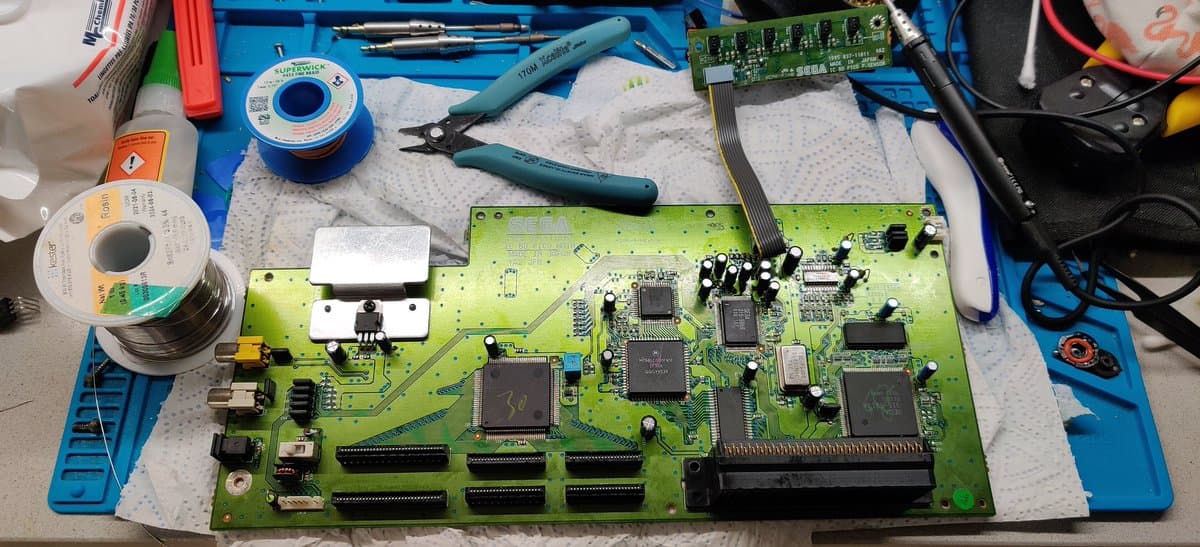 Sega Pico mainboard during recap repair, showing green PCB with ICs, capacitors, solder spool, desoldering tools, and multimeter on blue mat.