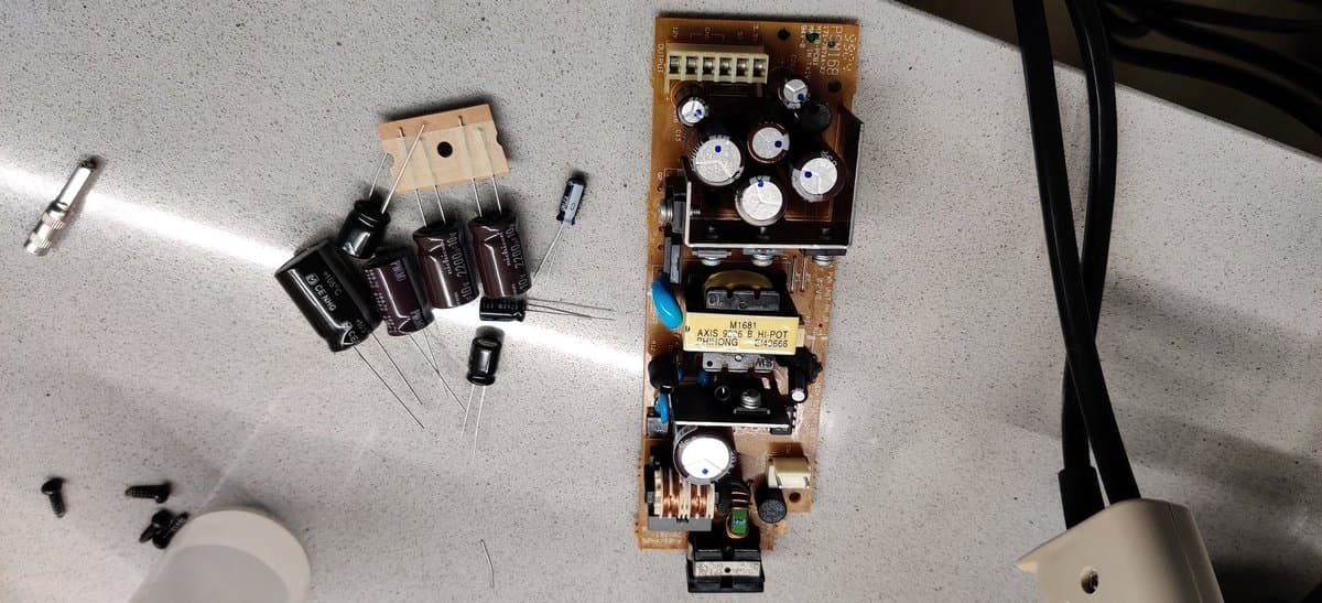 Disassembled Dreamcast VA0 PSU with capacitors removed, controller PCB with added fuse and battery mod components on cork board surface.