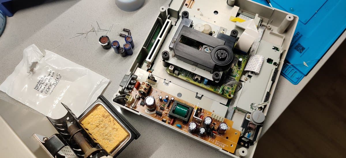 Disassembled Saturn Model 2 PSU showing recapped power board with electrolytic capacitors, inductor coil, and transformer alongside solder iron and replacement components.