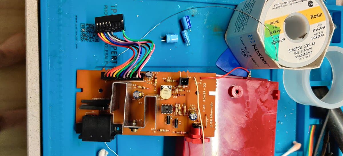 Famicom Disk System circuit board during recap with orange PCB, multicolored wiring harness, solder spool, and red capacitor housing visible.