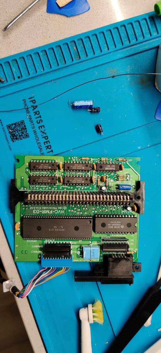 Famicom Disk System PCB during capacitor replacement with desoldered components and fresh solder joints visible on green circuit board.