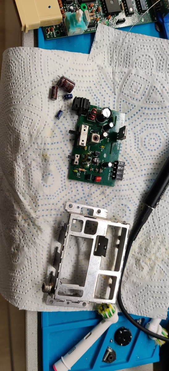 Famicom motherboard with new capacitors installed, composite mod board above, and white plastic housing below on workbench during repair and modding.