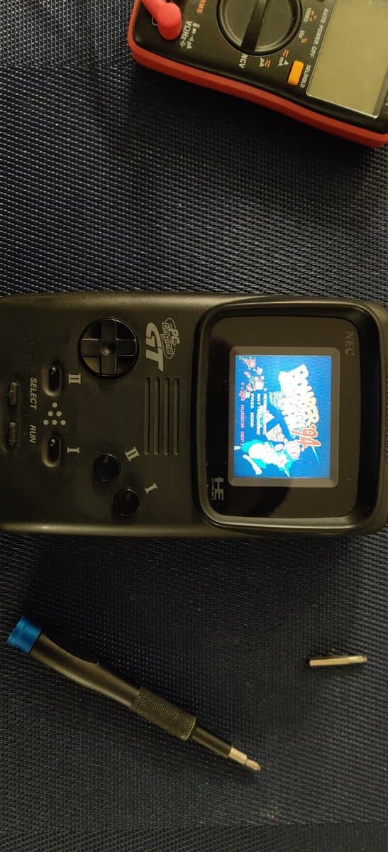 NEC PC Engine GT handheld with screen displaying Bomberman '94, alongside red multimeter and soldering iron on workbench during capacitor replacement repair.