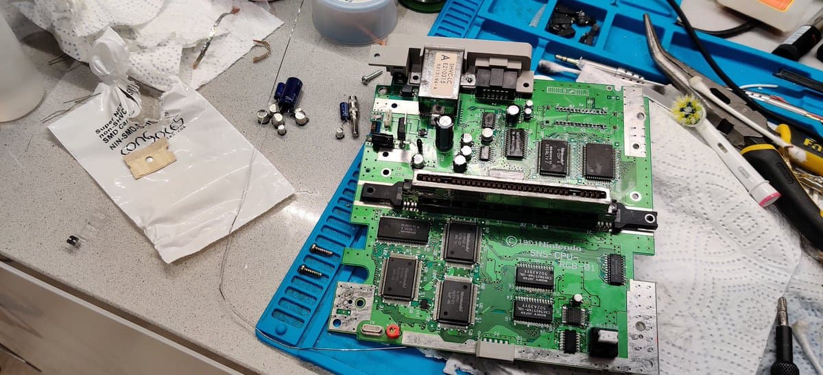Super Famicom main PCB on blue organizer with removed capacitors, diodes, and relay component during repair work on gray surface.