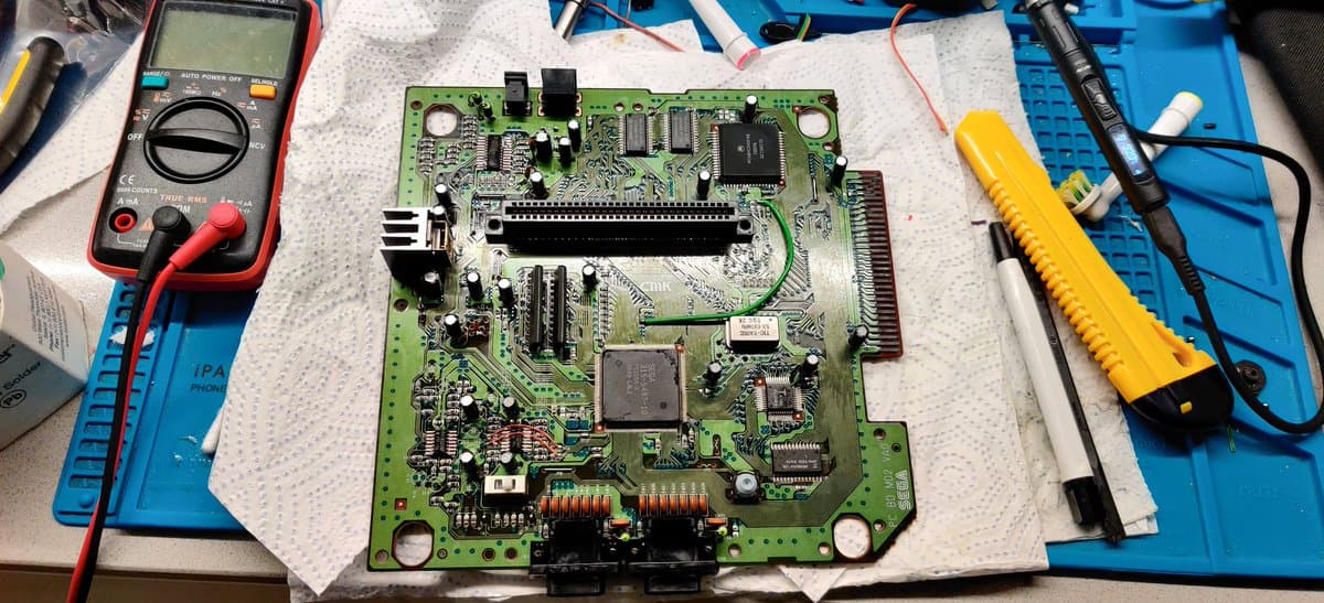Sega Mega Drive II motherboard undergoing repair with multimeter, soldering iron, and desoldering tool visible; capacitors and bypass work in progress.