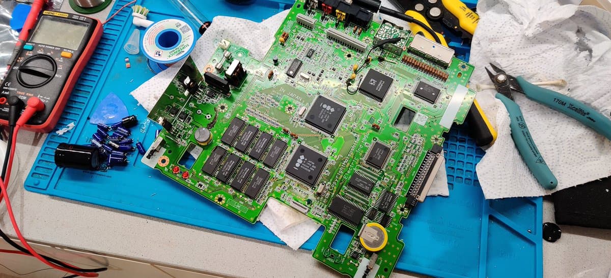 Panasonic FZ-1 motherboard undergoing repair with digital multimeter, solder, desoldering braid, and tools visible; multiple ICs and recapped board shown.