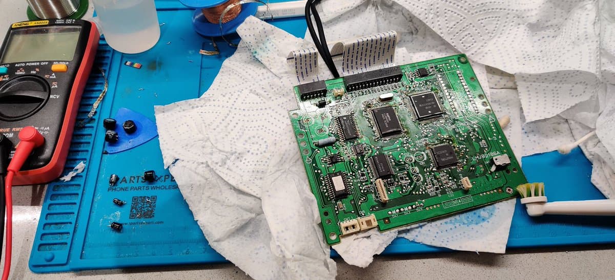 Panasonic FZ-1 optical drive PCB with new capacitors installed, multimeter and soldering equipment on blue workmat during restoration.