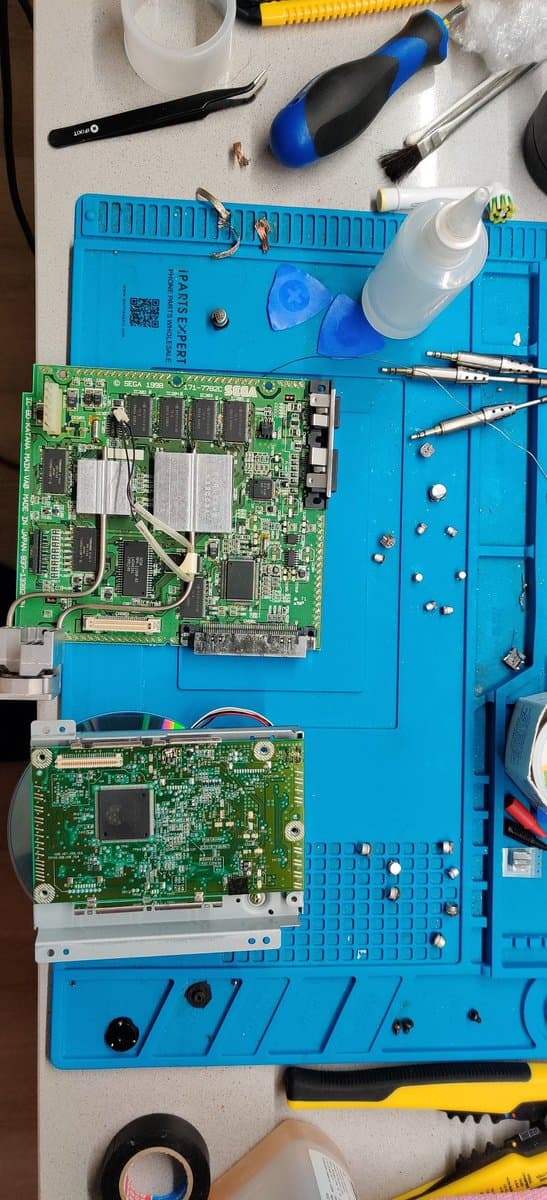 Dreamcast VA0 motherboard and optical drive PCB on blue mat during recap, surrounded by soldering tools, tweezers, and desoldering pump.