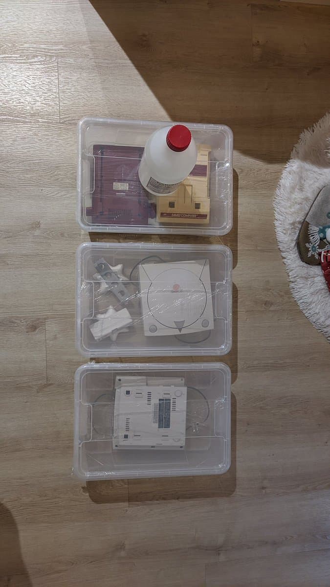 Three clear plastic containers displaying disassembled Dreamcast VA0 components: motherboard, power supply, and GDEMU mod kit with red power button on wooden floor.