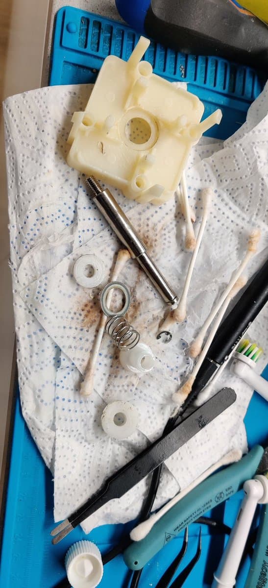 Dreamcast arcade stick joystick assembly disassembled on workbench: cream-colored plastic housing, metal shaft, springs, washers, and screwdriver for reassembly.