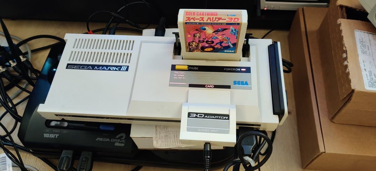 Sega Mark III with 3D Glasses adapter connected, Space Harrier 3D cartridge inserted, Mega Drive 16-bit console visible, repair setup on wooden surface.