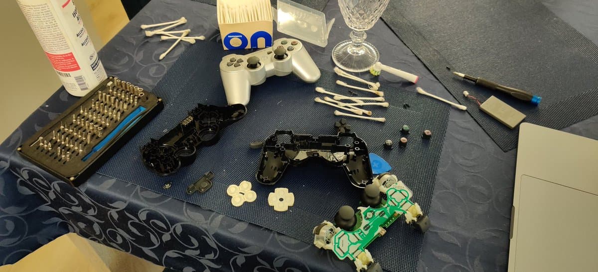Two PS3 controllers disassembled on workbench: black controller with shell off revealing circuit board and components; white controller partially disassembled. Cleaning tools, small parts, and supplies visible.