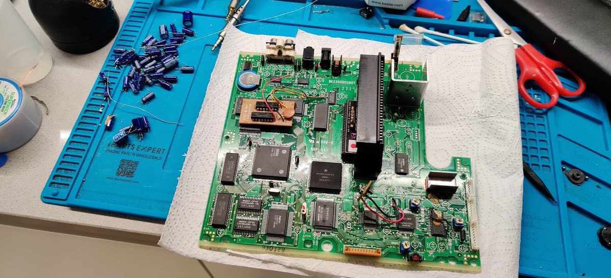 Sega Mega-CD 2 mainboard undergoing recapping repair, with desoldered capacitors scattered on blue workmat and fresh solder joints visible.