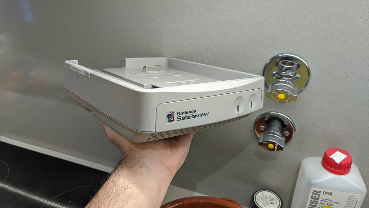 Nintendo SatellaView console held in hand after retrobrighting, shell displaying improved whiteness; various electronic components visible nearby on workbench.